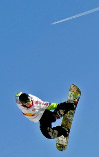 Un snowboardeur avec un haut blanc et rouge et un pantalon noir effectuant un big air. Un avion est pr&eacute;sent arri&egrave;re plan dans le ciel.